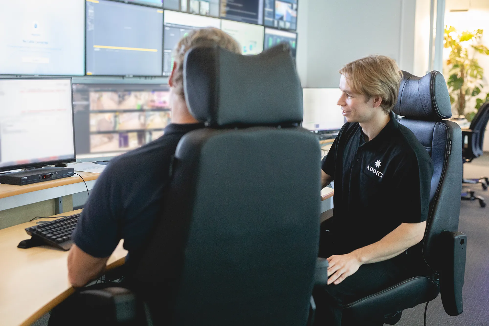 två Addicimedarbetare framför en vägg av skärmar på larmcentralen